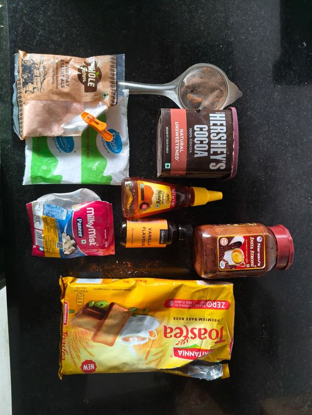 Flat lay of ingredients for paneer tiramisu: paneer, toned milk, honey, vanilla essence, pink salt, instant coffee, unsweetened cocoa powder, and Britannia Toastea rusks.