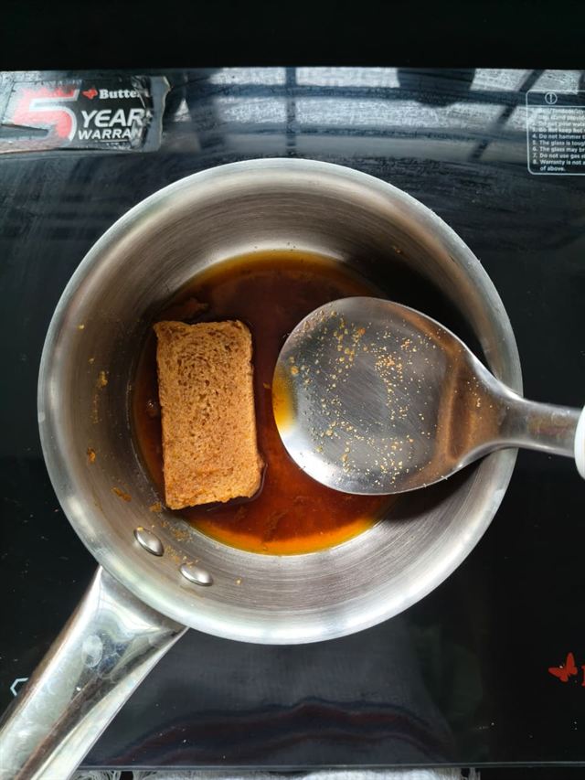 Overhead shot of a small saucepan with coffee syrup and a soaking rusk piece, spoon resting inside, on an induction stovetop.